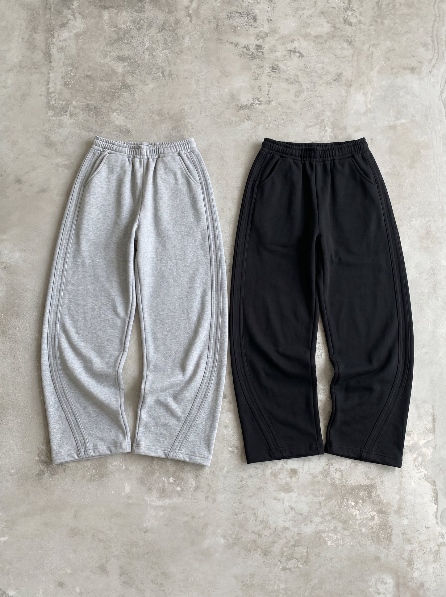 グレーとブラックのワイドスウェットパンツ2色の平置き。前面に脚のラインに沿ったカーブの切り替えステッチ。ワイドシルエットでドローコードウエスト。